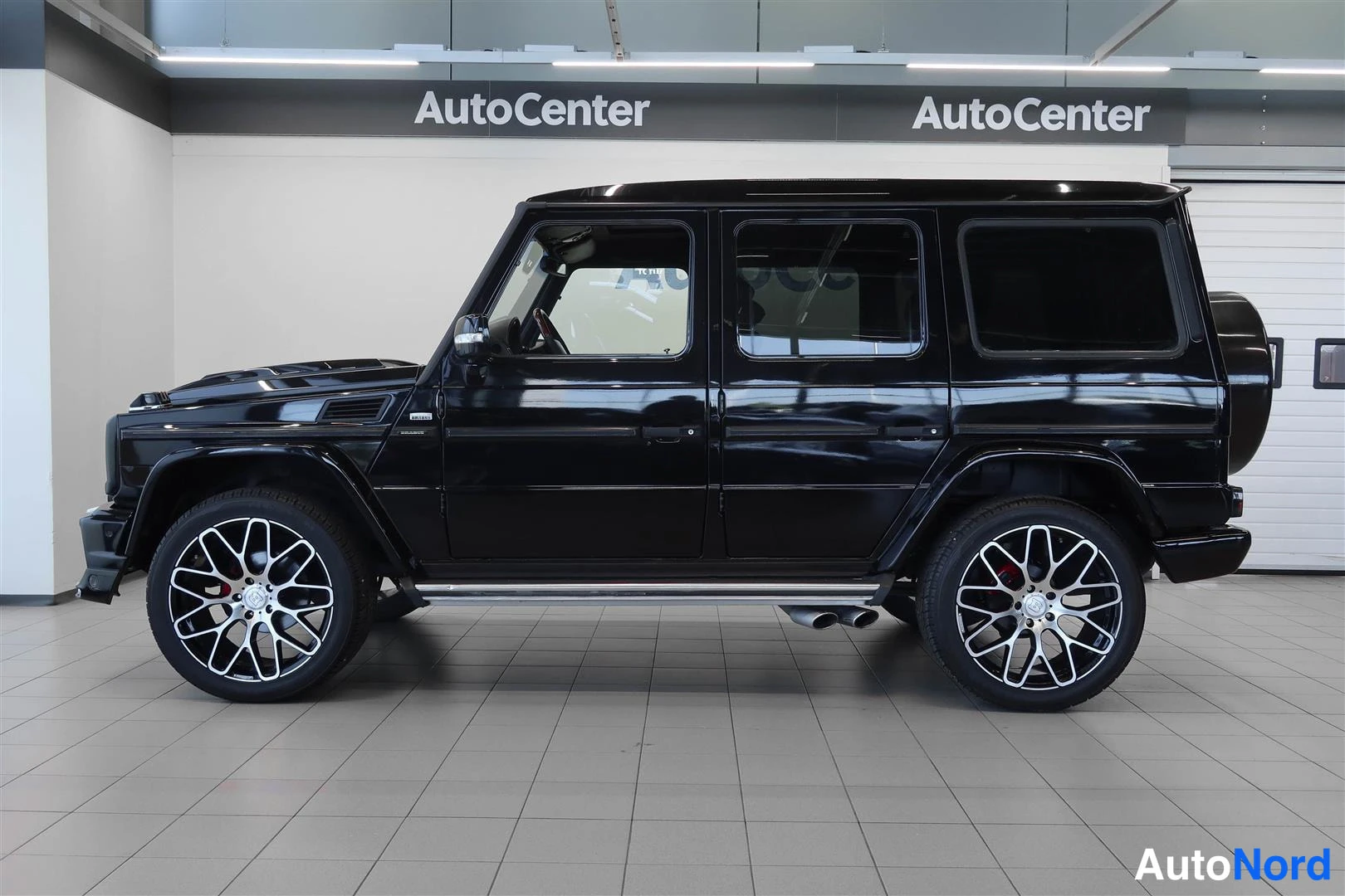 Sivulta: Musta Mercedes-Benz G -maasturi toiselta sivulta kuvattuna sisätiloissa, hopeat kevytmetallivanteet.