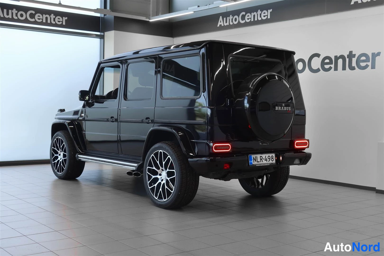 Takaviisto: Musta Mercedes-Benz G takaviistosta sisätiloissa; perä ja vasen kylki näkyvissä, varapyörä kannella ja rekisterikilpi.