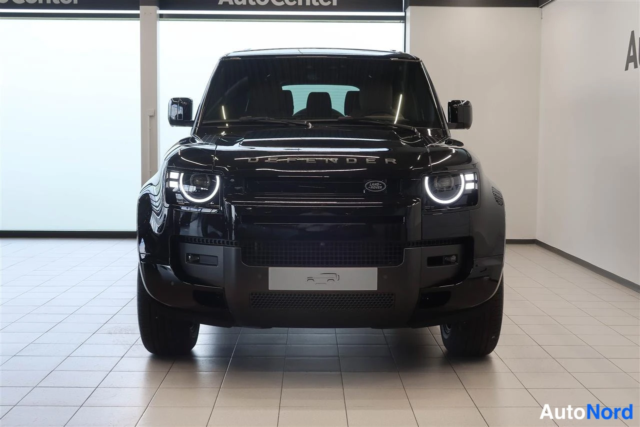 Edestä: Land Rover Defenderin etunäkymä edestäpäin, pyöreät LED-päiväänajovalot ja Land Rover -merkki konepellissä, Autocenter-tilassa.