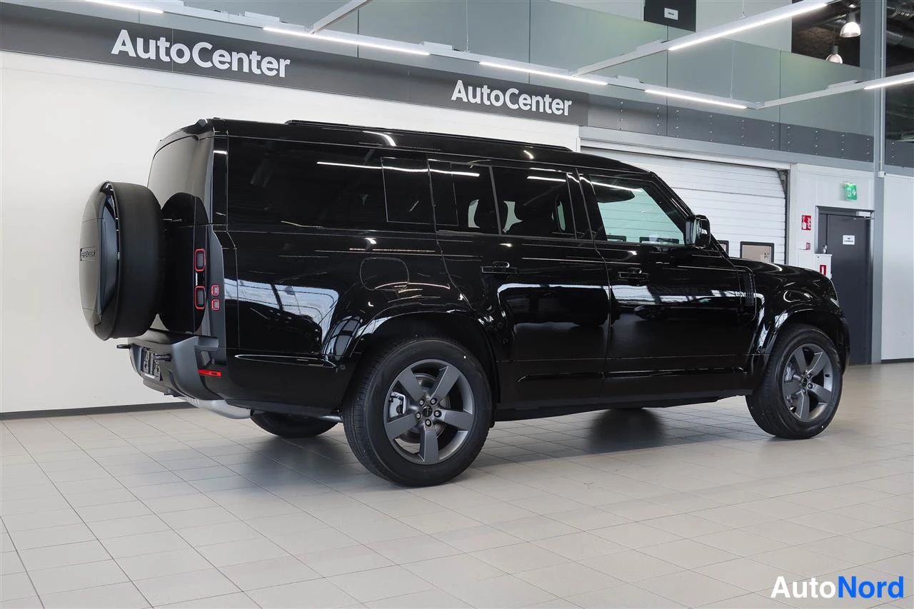 Takaviisto: Land Rover Defenderin takaosa kolmannelta kuvakulmalta; musta koripaneeli, takavalojen elementit ja takana kiinnitetty varapyörä näkyvät showroomissa.