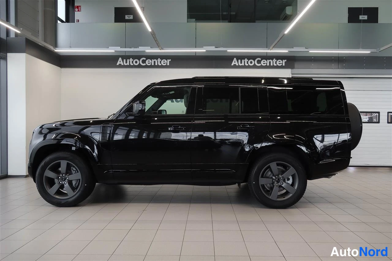 Sivulta: Land Rover Defenderin sivuprofiili; musta koripaneeli, kattokaiteet ja tummat kevytmetallivanteet näkyvät showroomin valkolattialla.
