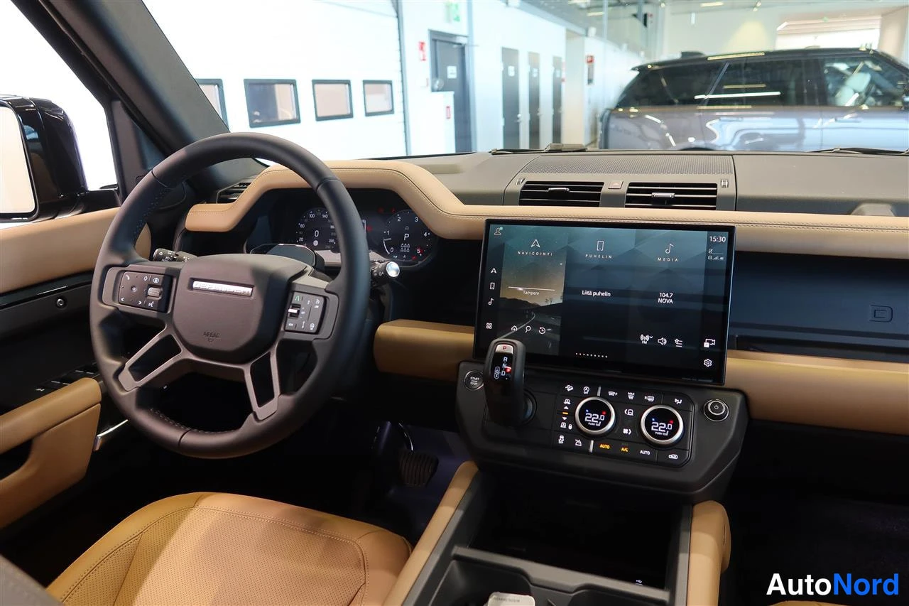 Muu: Land Rover Defenderin ohjaamo: tumma nahkaohjauspyörä, analogiset mittarit, keskiöön asennettu suuri infotainment-näyttö (radiotoiminto, 15:30 näkyy), automaatt
