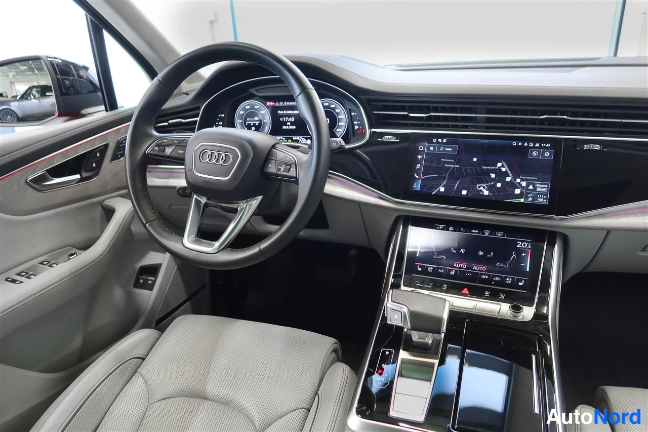 Muu: Audi Q7:n ohjaamo: Audi-merkkinen ohjauspyörä, digitaalinen mittaristo, kaksi kosketusnäyttöä keskikonsolissa ja harmaat nahkaistuimet.