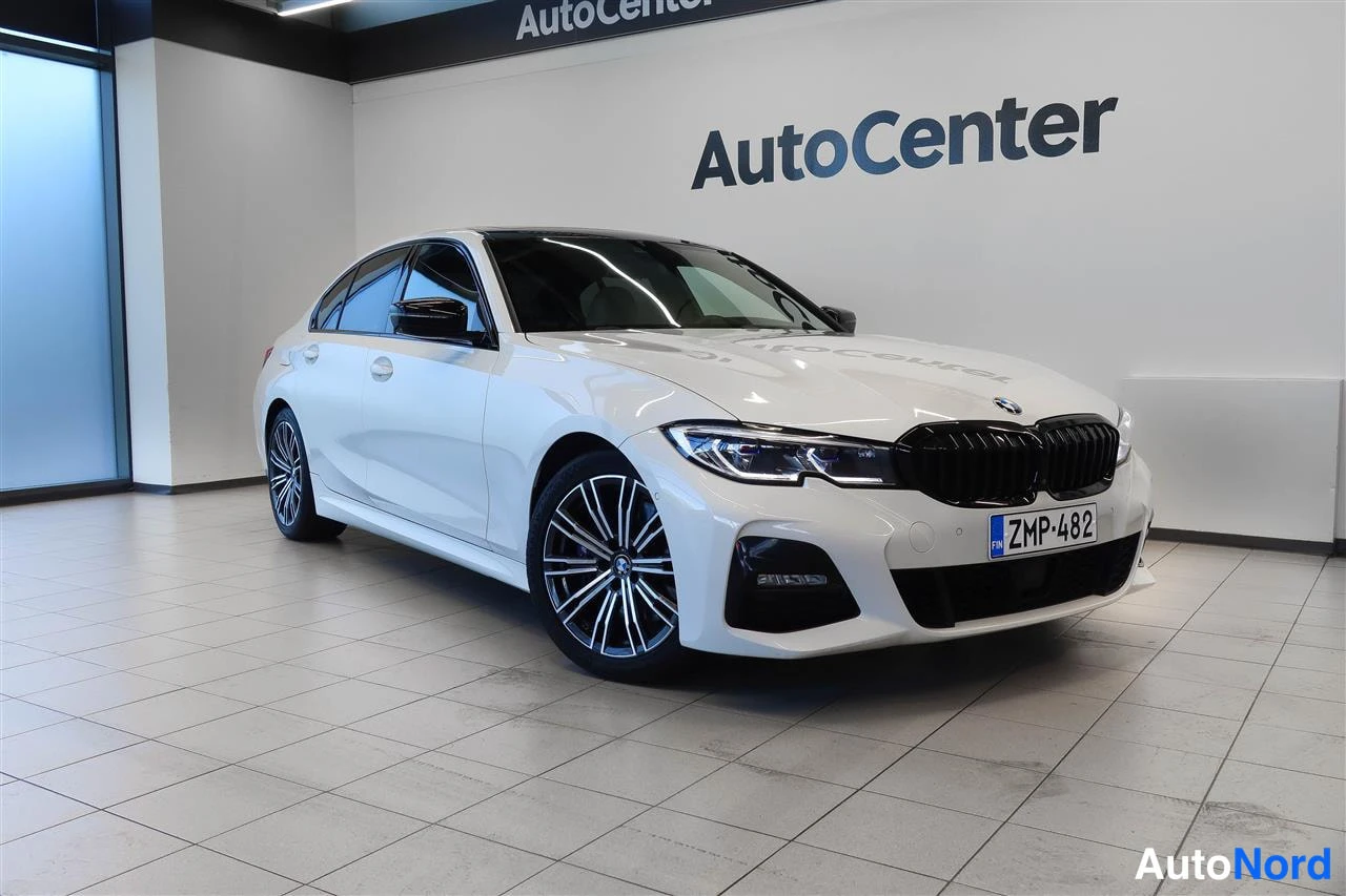 Etuviisto, nokka oikealle: Valkoinen BMW 330 -sedan kulmaetuvasivukuva autonhallissa, musta etusäleikkö ja -kilpi.