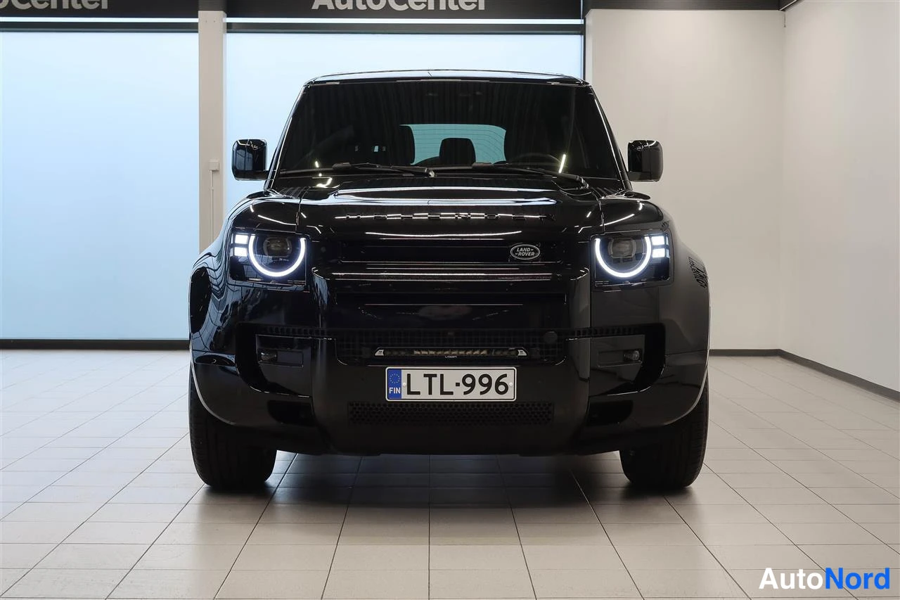 Edestä: Musta Land Rover Defender suoraan edestä kuvattuna, pyöreät LED-päiväajovalot ja rekisterikilpi.