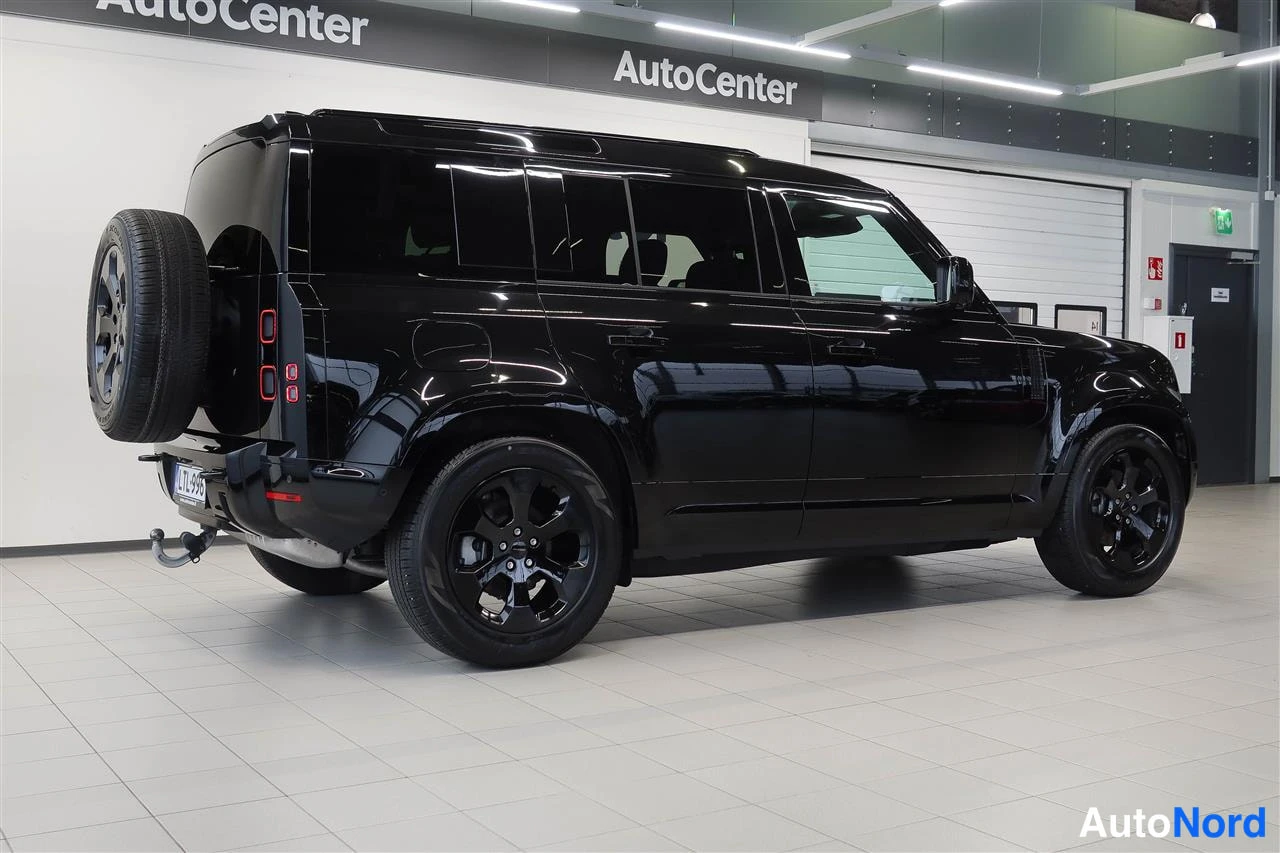 Takaviisto: Mustan Land Rover Defenderin takaosa ja vasen sivu autotallissa, varapyörä takaluukissa.
