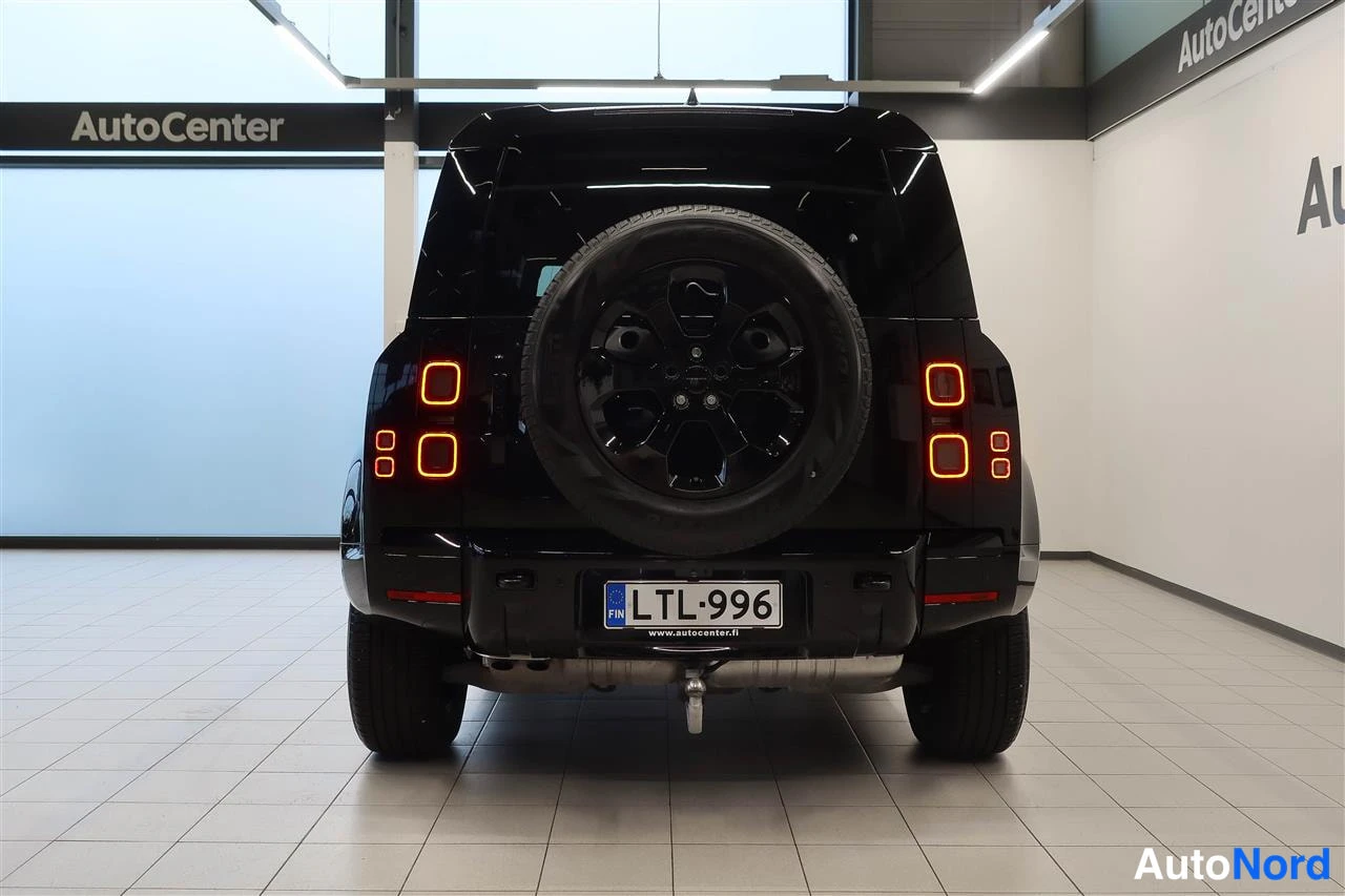 Takaa: Mustan maastoa auton takaosa suoraan takaa; varaheitin kannessa, punaiset takavalot palasivat, rekisteritunnus ja valkoinenAutoCenter-hallissa.