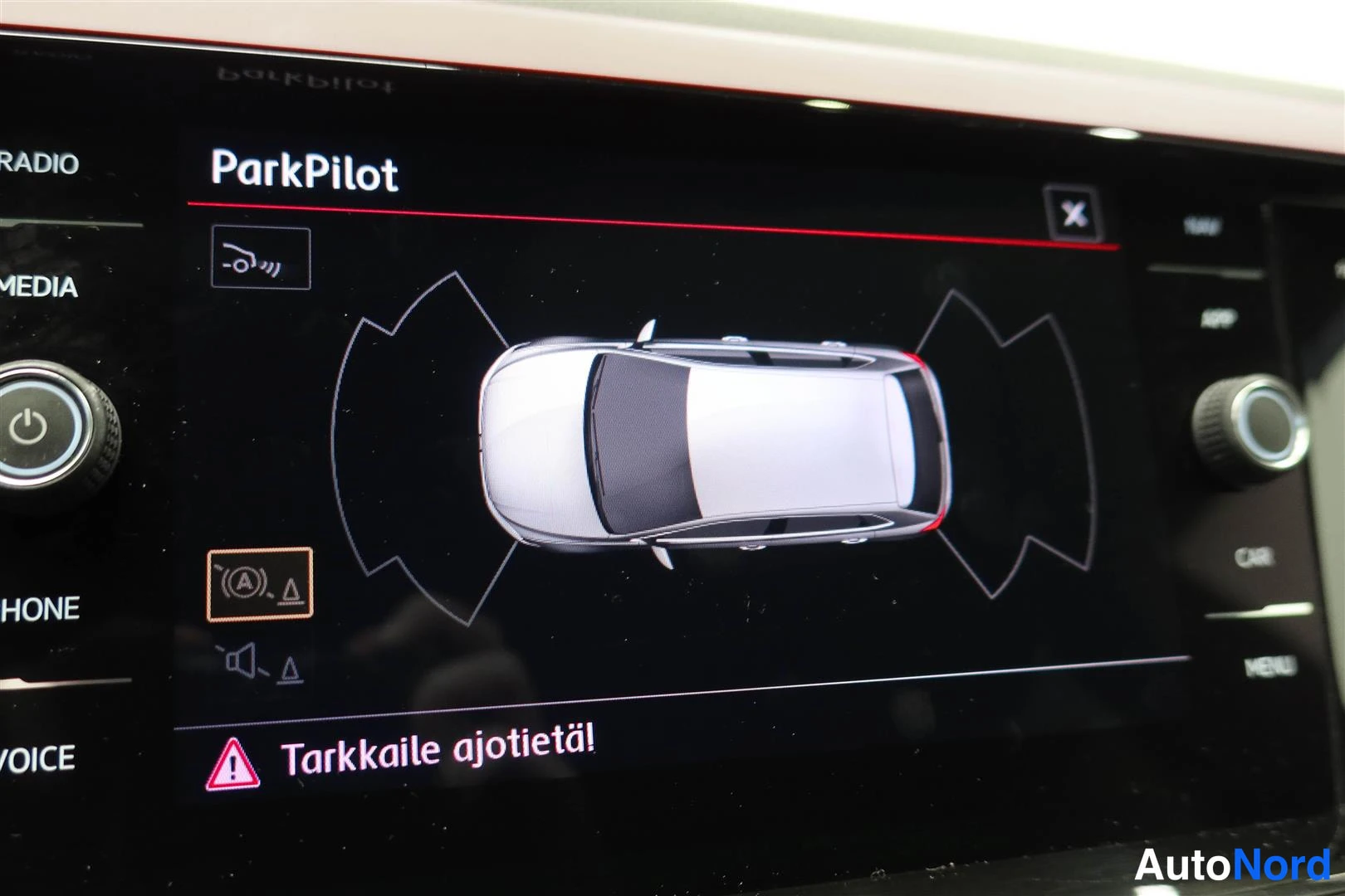 Muu: Auton radio-/infotainment-näyttö, jossa on ParkPilot-tutkatilanne: ajoneuvo valkoisena ylhäältäpäin näkymässä ja Tarkkaile ajotietä! -varoitusviesti.