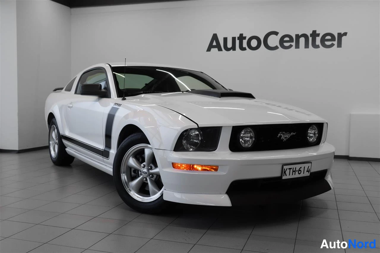 Etuviisto, nokka oikealle: Valkoinen Ford Mustang etukulmasta, konepelti ja etupuskuri näkyvät, musta bossesohjattu raita ovella, rekisterikilpi.