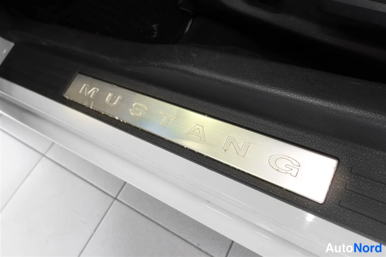 Yksityiskohta: Lähikuva oven kynnyksen metallilistasta, johon on kaiverrettu teksti MUSTANG; auton kynnyksen osa valkoinen.