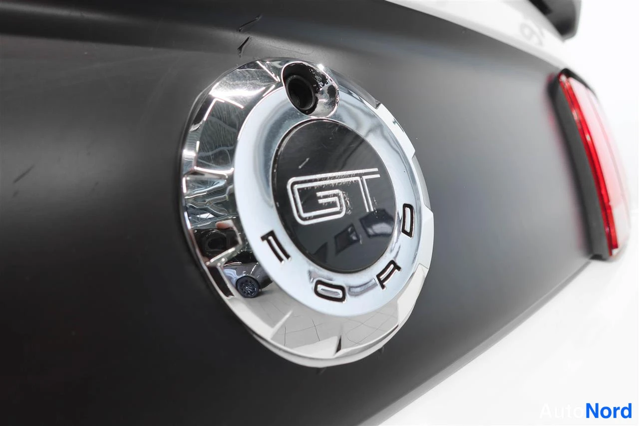 Takaa: Mustan autonpinnan lähikuva: kiiltävä kirkas pyöreä tankkiluukun kansikorkki, jonka keskellä musta kehys ja hopeinen GT-logo ja alapuolella FORD-teksti. Oikeall