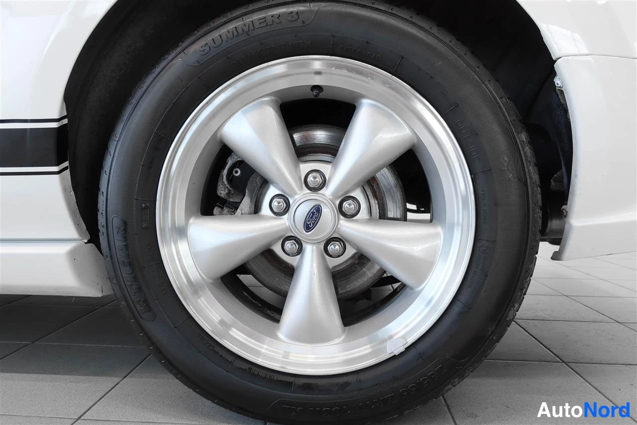 Sivulta: Valkoisen auton takapyörän lähikuva: kirkashopea moniSWEP-pölykoppa, keskeinen Ford-logo, pyöreä keskus. Mustan kumin pinnassa tekstiä 'SUMMER 3'. Taustassa val