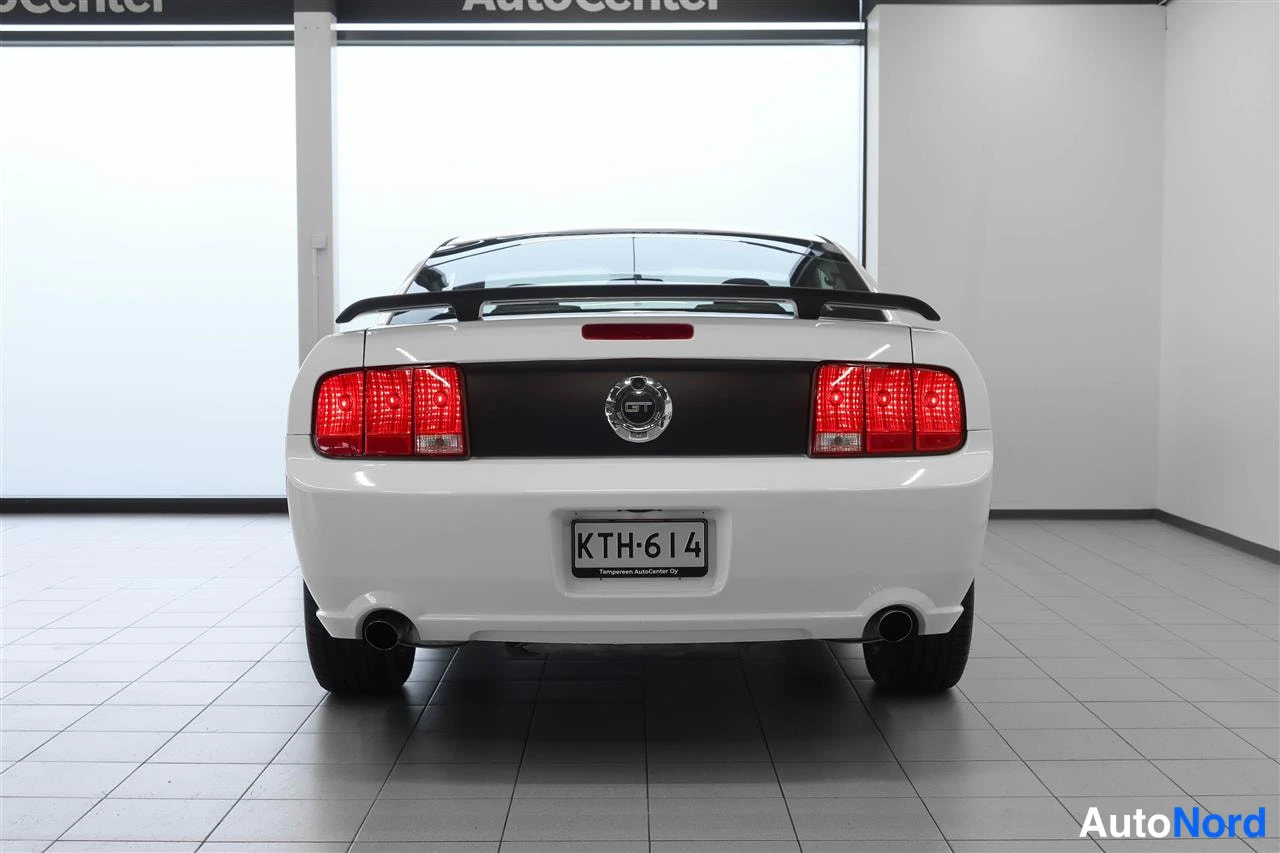 Takaa: Valkoisen Ford Mustangin takaosa näkyvissä suoraan takaa; punaiset takavalot, musta spoileri, GT-emblem ja rekisterikilpi.