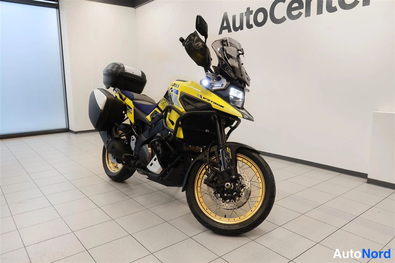 Etuviisto, nokka oikealle: Keltamusta Suzuki V-Strom 1050 -moottoripyörä etukulmasta oikealta, kultaiset vanteet, matkalaukku ja sivulaukut, sisätiloissa.