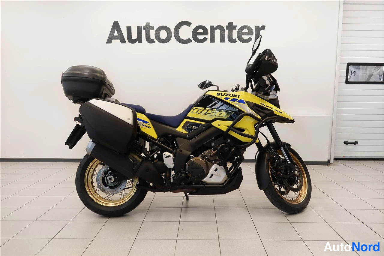 Sivulta: Suzuki V-Strom 1050 sivuprofiilista, 1050-kylki, kelta-mustat korit, matkalaukku ja sivulaukut, kultaiset vanteet sisätiloissa.