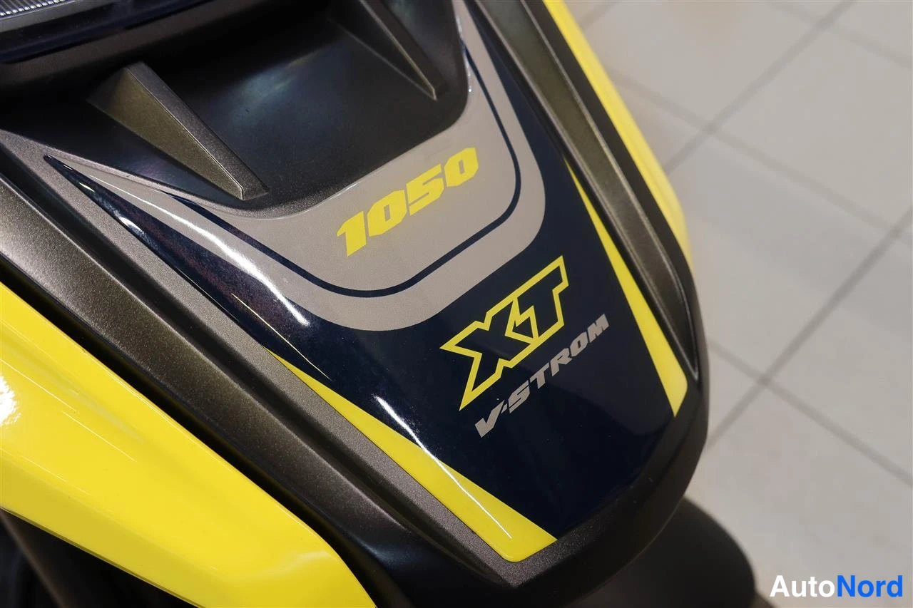 Yksityiskohta: Lähikuva keltamustan Suzuki V-Strom 1050 XT -moottoripyörän keulakilvestä, jossa lukee 1050, XT ja V-STROM.