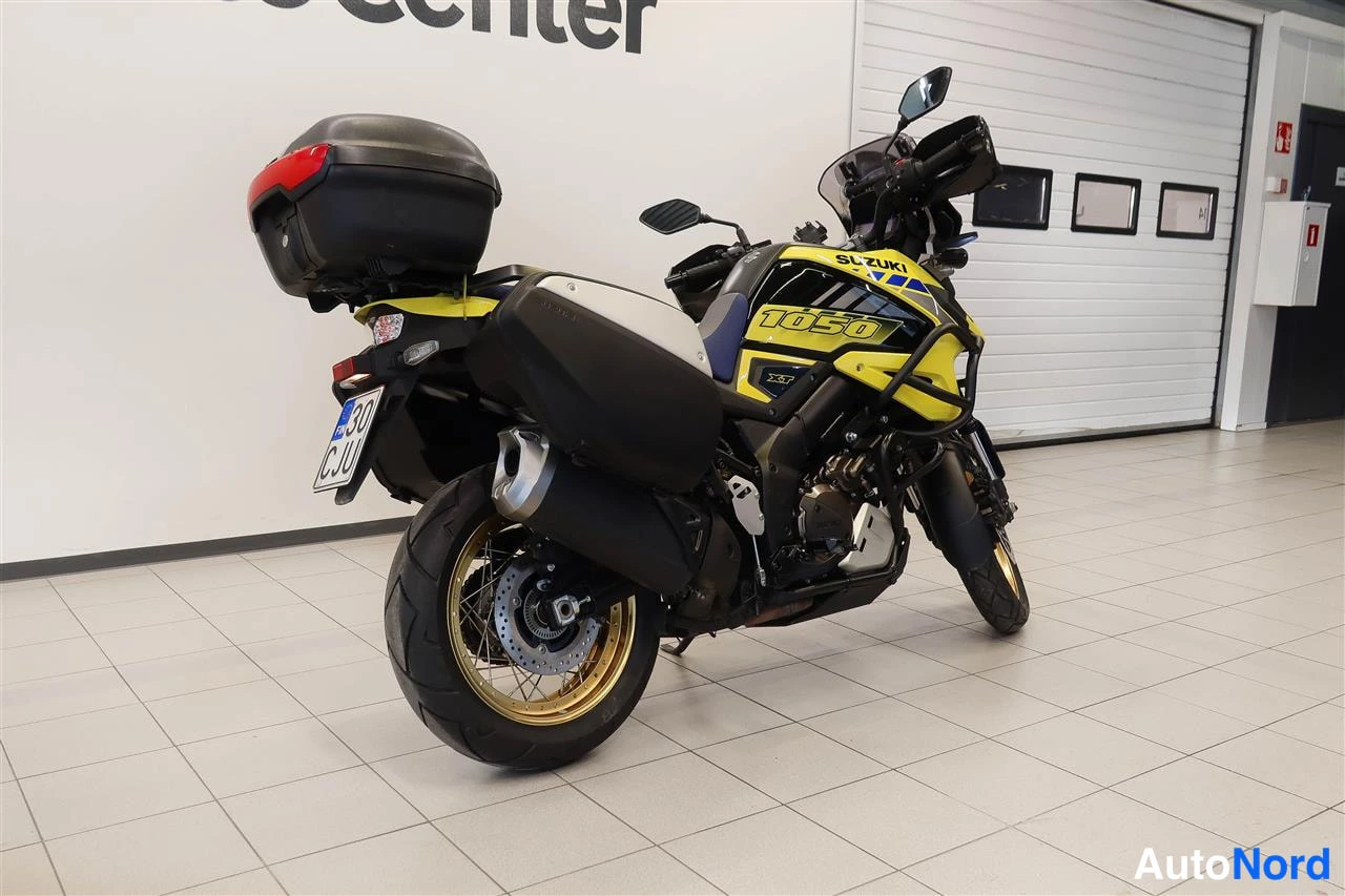 Takaviisto: Suzuki V-Strom 1050 takakulmasta vasemmalta, takalokasuoja ja pakoputki, musta matkalaukku, rekisterikilpi, sisätiloissa.