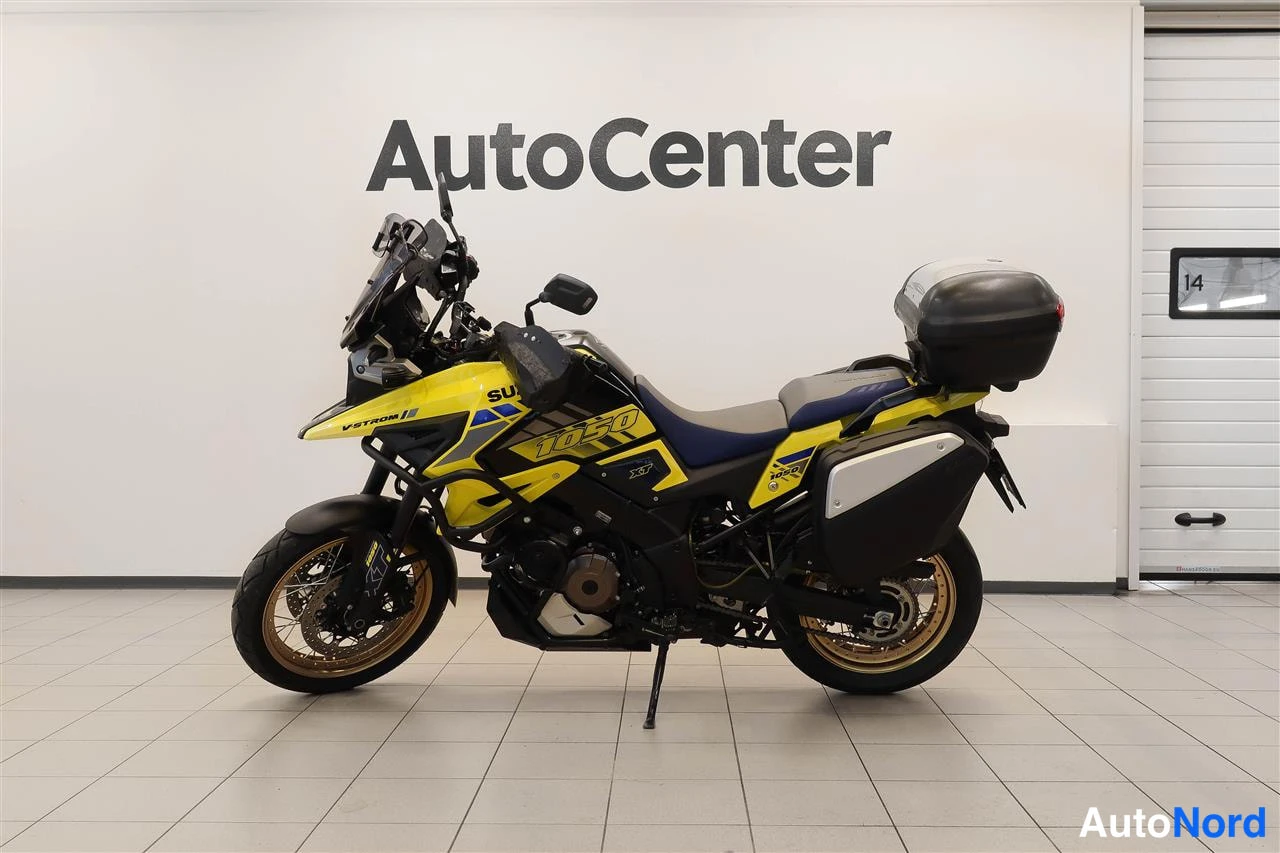 Sivulta: Suzuki V-Strom 1050 -moottoripyörä sisätilassa sivuprofiilista, valkoisen seinän ja AutoCenter-kyltin edessä.