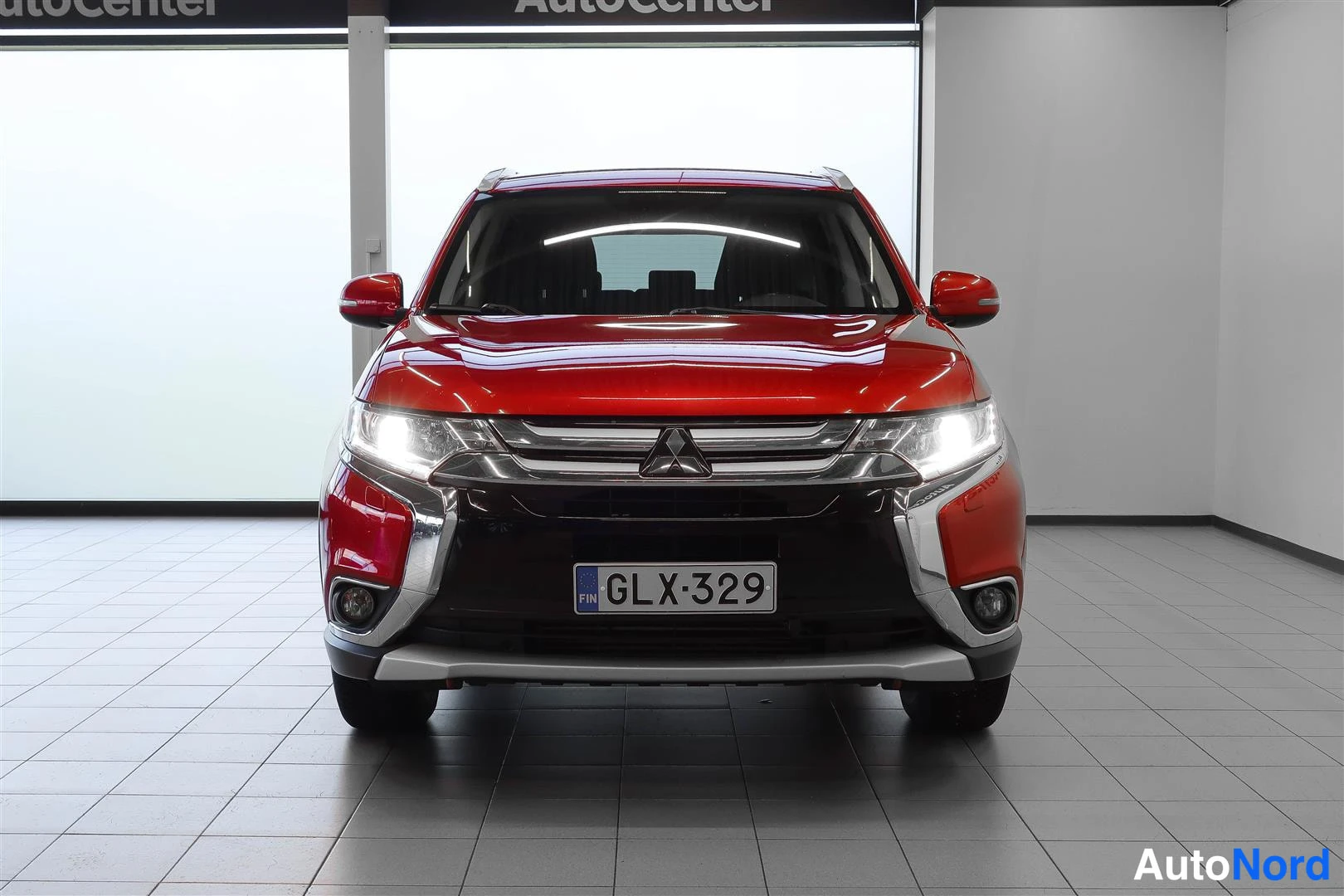 Edestä: Etukuva punaisesta Mitsubishi Outlanderista sisätiloissa, ajovalot päällä ja rekisteritunnus.