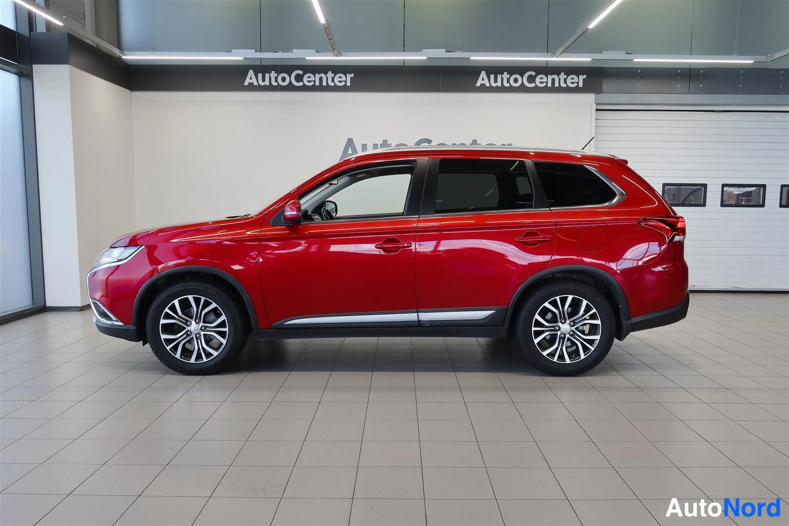 Sivulta: Punainen Mitsubishi Outlander kokonaan sivusuunnassa kuvattuna autoluupissa; molemmat sivut, kattokaiteet ja alumiinivanteet näkyvät.