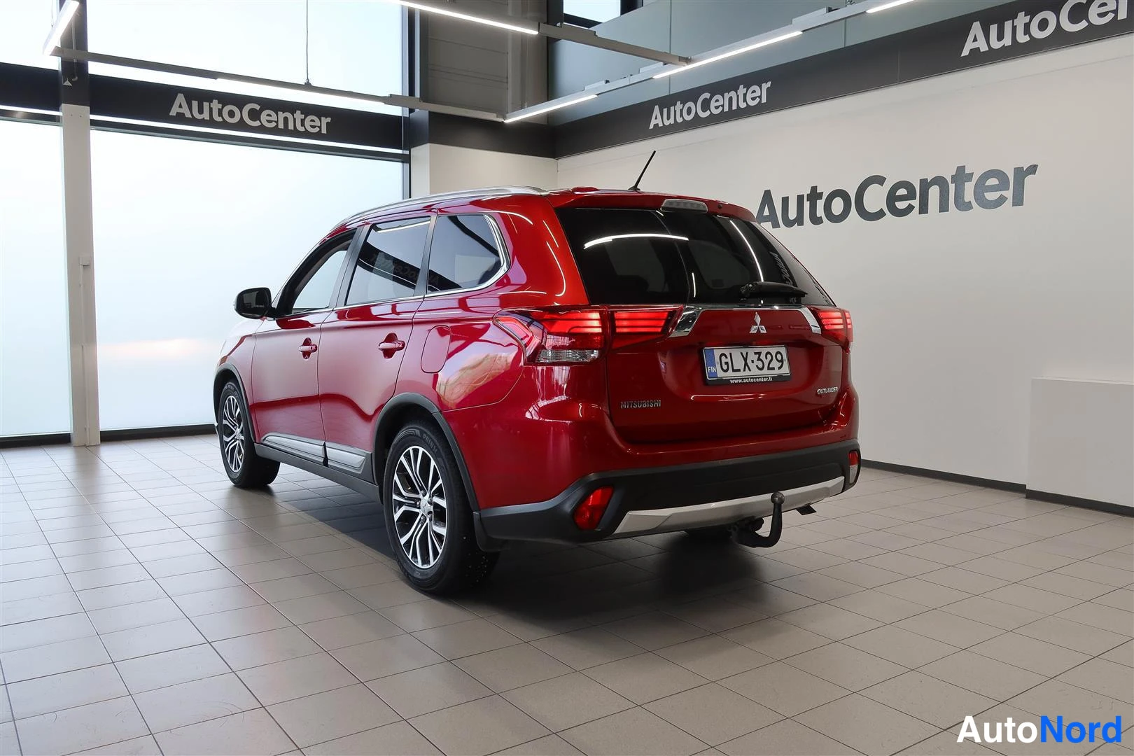 Takaviisto: Punainen Mitsubishi Outlander takaa ja vasenta kylkeä kuvattuna autoluupissa; rekisterikilpi, takavalojen poikittaisrakenne ja vetokoukun kaula näkyvät.