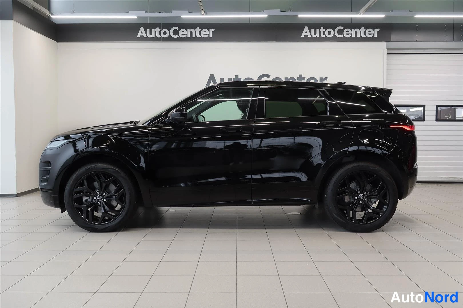 Etuviisto, nokka vasemmalle: Land Rover Range Rover Evoque 2023, musta, lähes sivukuva oikealta; ovet ja mustat vanteet näkyvissä AutoCenter-hallissa.