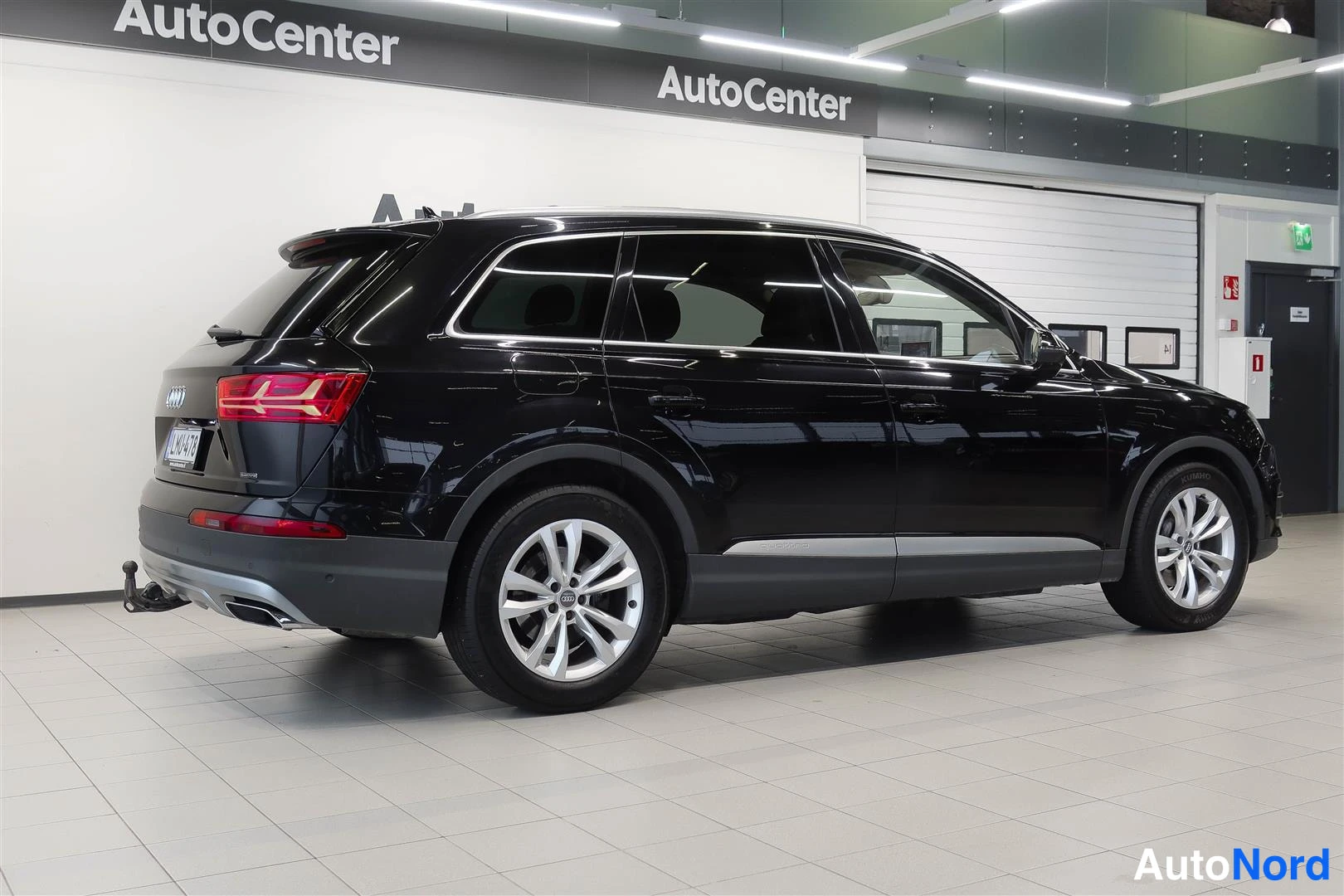 Takaviisto: Musta Audi Q7 katumaasturi takana vasemmalta kuvattunaAutocenter-hallissa, valmiiksi asennettu vetokoukku ja kaksoispakoputki näkyvät.