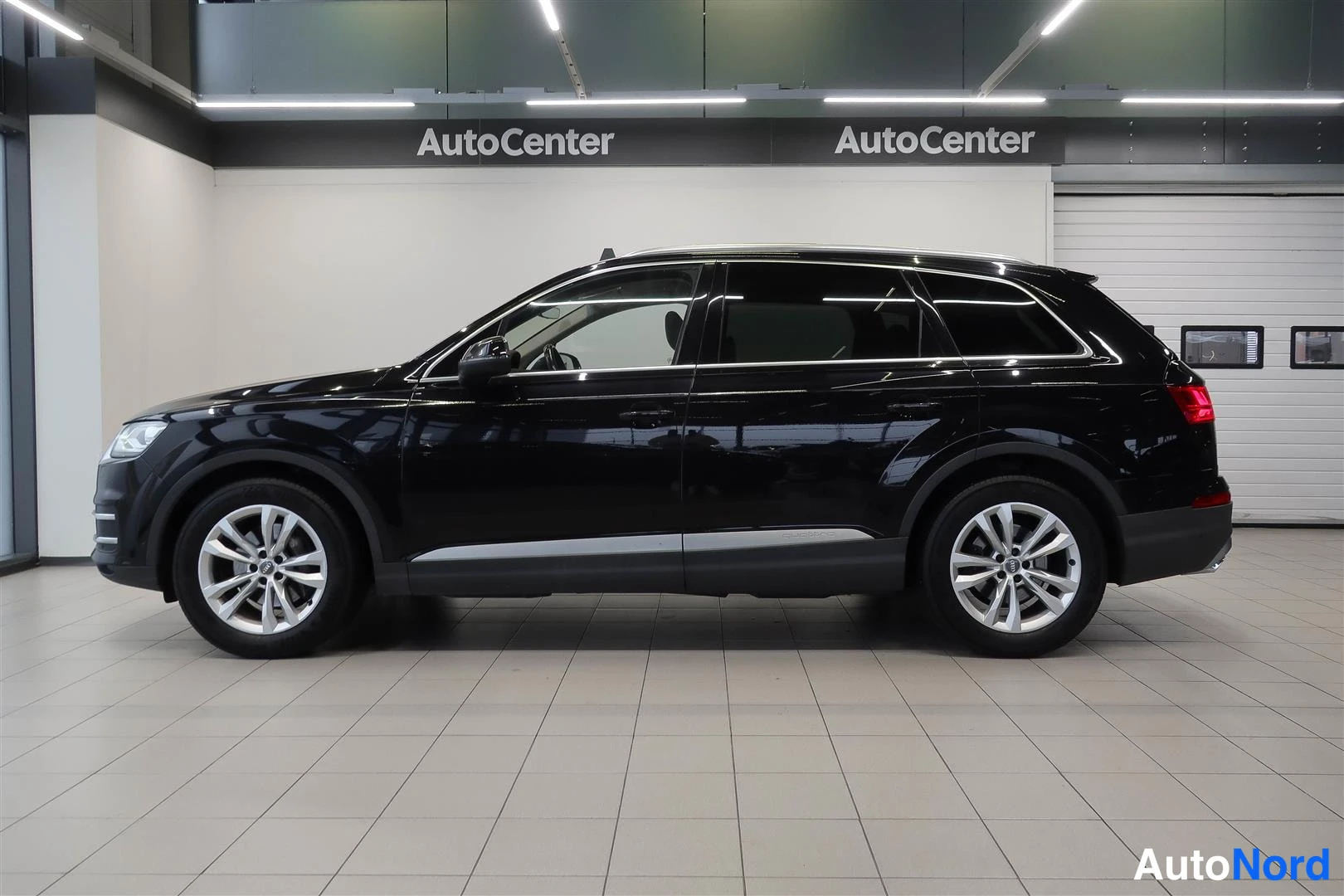 Sivulta: Musta Audi Q7 katumaasturin kylki täsmällisesti sivulta kuvattunaAutocenter-hallissa, hienot renkaat ja kromilista.