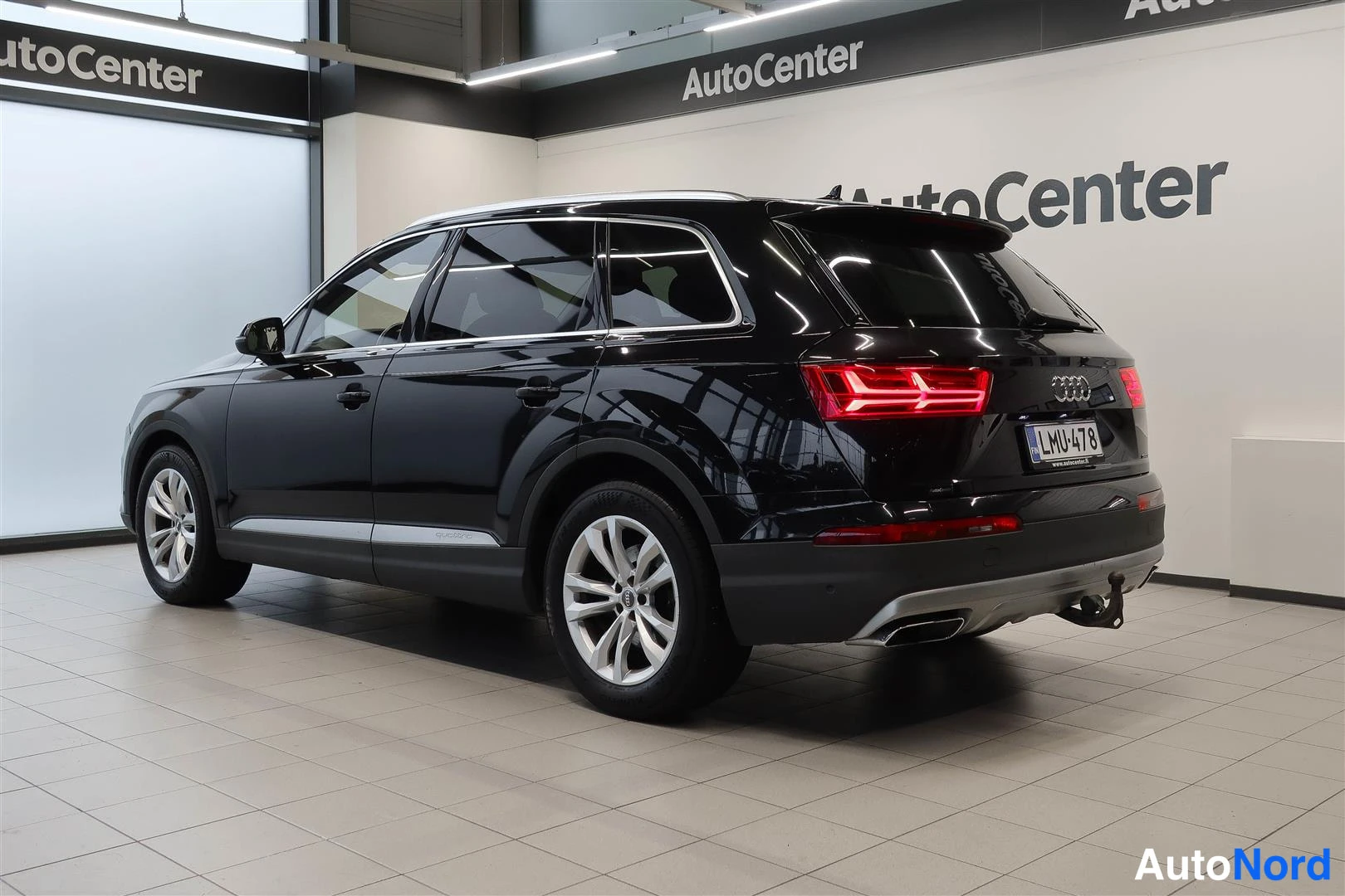 Takaviisto: Musta Audi Q7 katumaasturi takaa vasemmalta kuvattunaAutocenter-hallissa, rekisteri ja vetokoukku näkyvät, takaosa lampor punaisena.