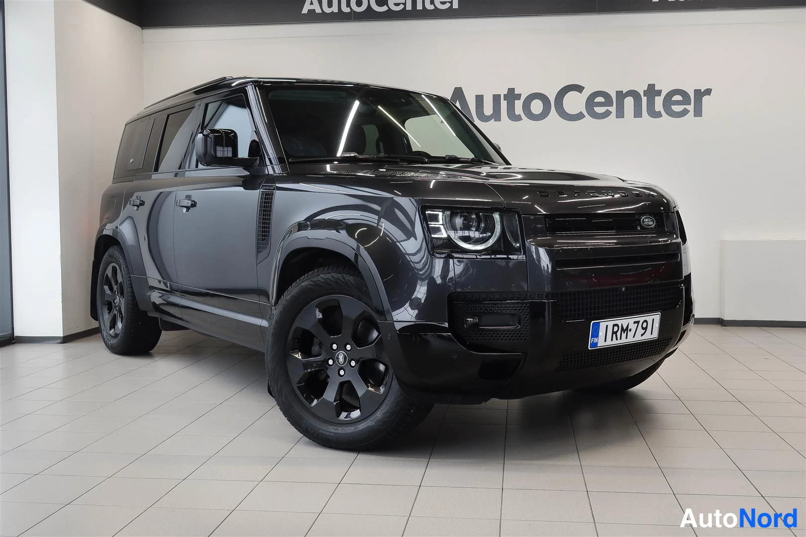 Etuviisto, nokka oikealle: Land Rover Defender 110 -maast-auto etukulmasta oikealta, tummanharmaa korinen ajoneuvo AutoCenter-näyttelytilassa, rekisteritunnus RM-791.