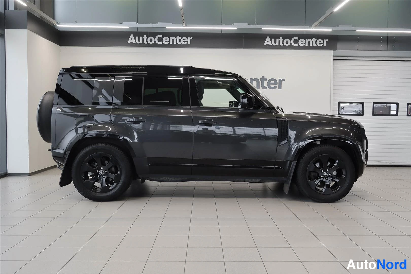 Sivulta: Land Rover Defender 110 -maast-auto sivulta kuvattuna vasemmalta, tummanharmaa kori, mustat kiiltävät vanteet, takana varapyörä.