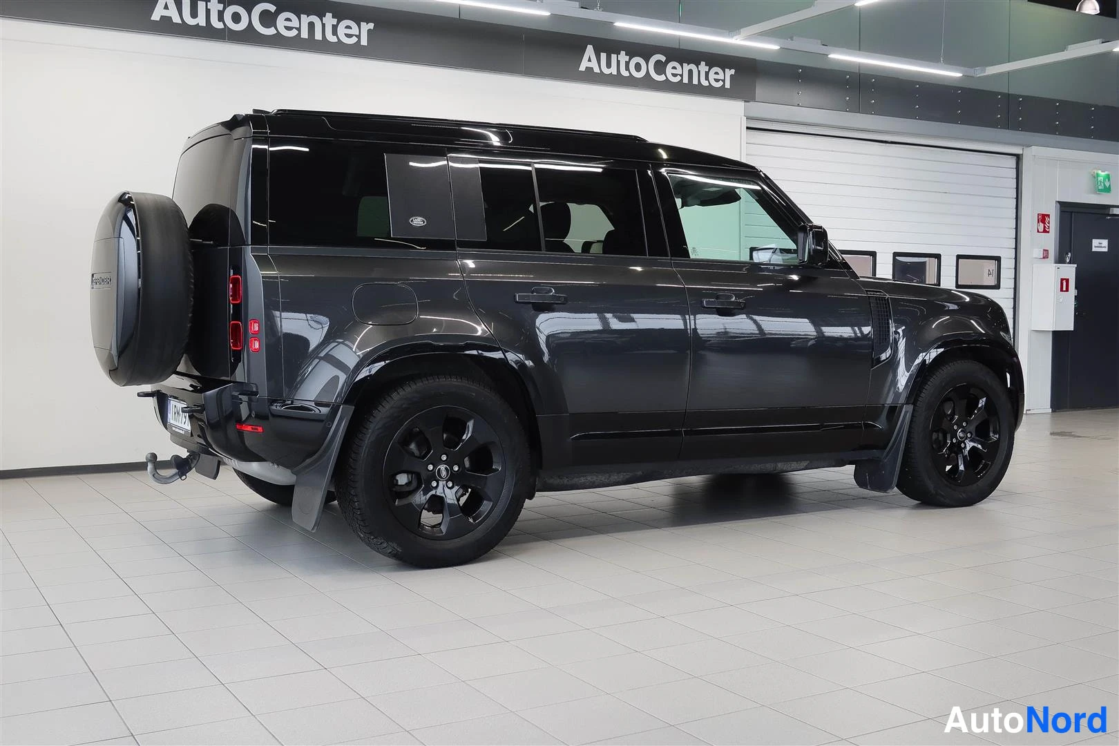 Takaviisto: Tummanharmaan Land Rover Defenderin takakulman sivunäkyvissä auton hallissa, taakse asennettu varaova ja mustat kevytmetallivanteet.
