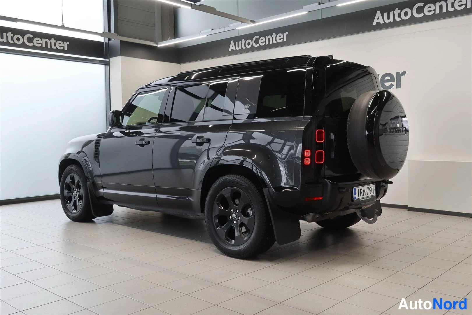 Takaviisto: Tummanharmaan Land Rover Defenderin takakulma sivulta kuvattuna hallissa; takavalojen punaiset osat ja rekisterikilpi näkyvät.
