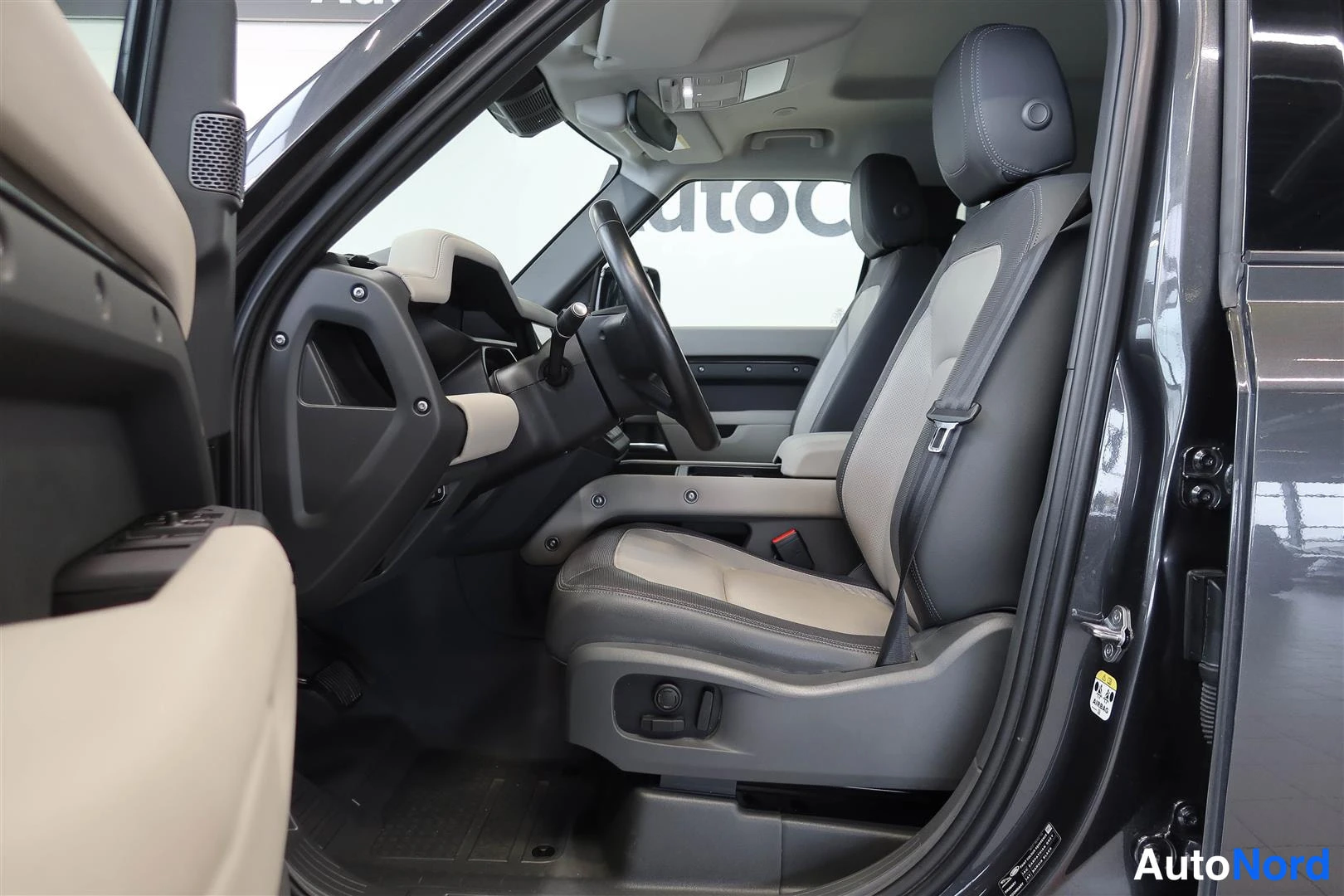 Muu: Land Rover Defenderin sisätilat: kuskinistuin, ohjauspyörä ja kojelaudan vasen osa etupyyhkijöineen.