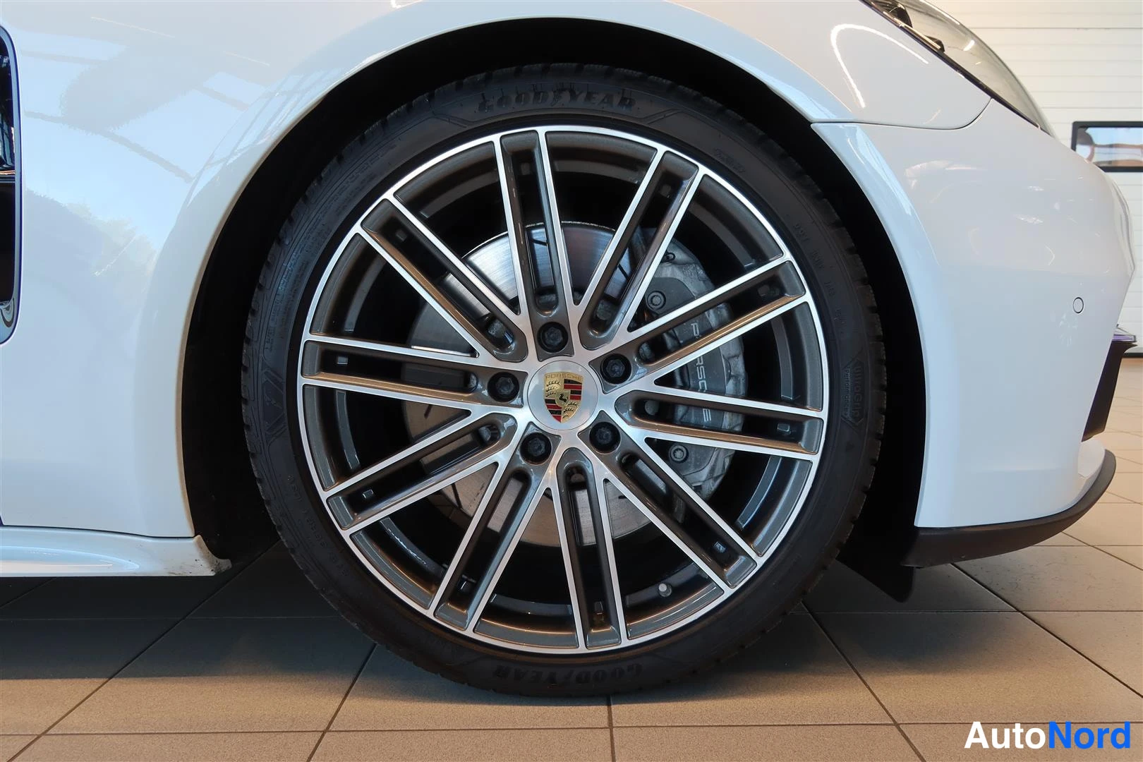Sivulta: Sivutärkeä kuva valkoisen auton etupyörästä: monisakarainen harmaa alumiinivanne, Porsche-merkki napassa, punainen jarrusatula ja Goodyear-rengas.