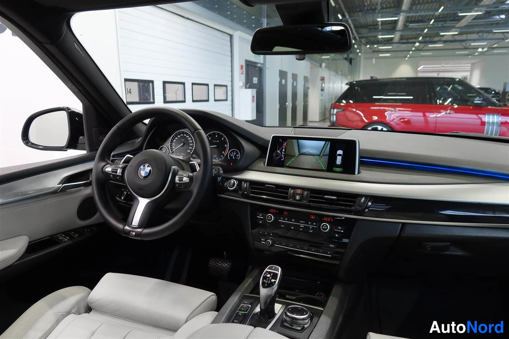 Muu: Ohjaamon näkymä kuljettajan puolelta: BMW-ohjauspyörä, mittaristo, keskinäytön kameraunnäkymä ja tumma kojelauta vaaleilla istuimilla.