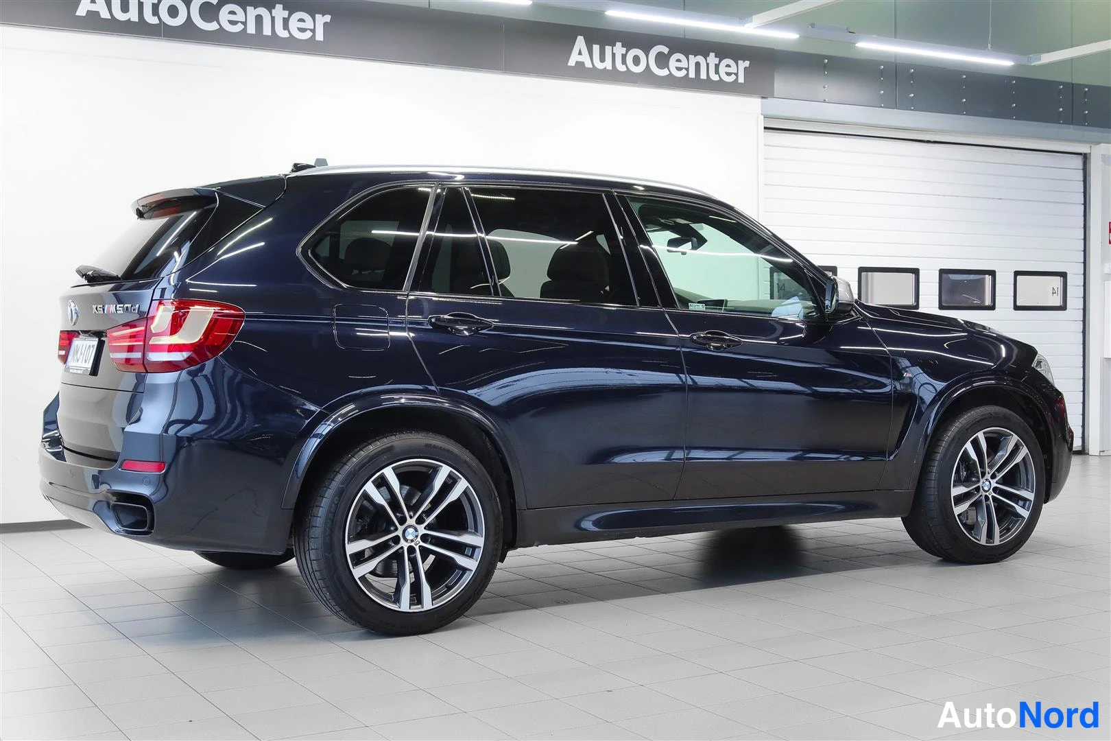 Takaviisto: Tummansinisen BMW X5:n takakulmanäkymä vasemmalta, takavalot ja rekisterikilpi näkyvät showroomissa.
