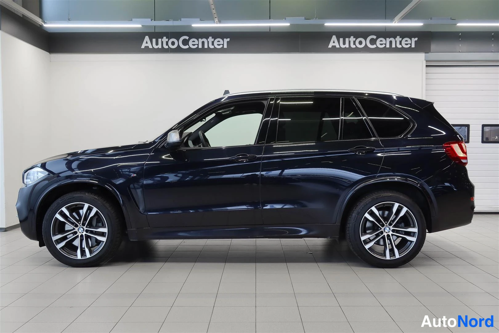 Sivulta: Tummansinisen BMW X5:n sivunäkymä vasemmalta, kattokaaret ja 20 tuuman metallivanteet näkyvät sisätiloissa.