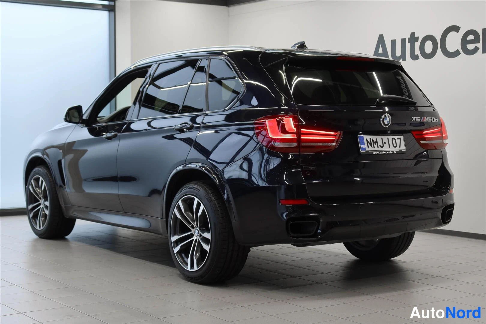 Takaviisto: Tummansinisen BMW X5:n takakulmanäkymä vasemmalta, takavalot ja rekisterikilpi näkyvät showroomissa.