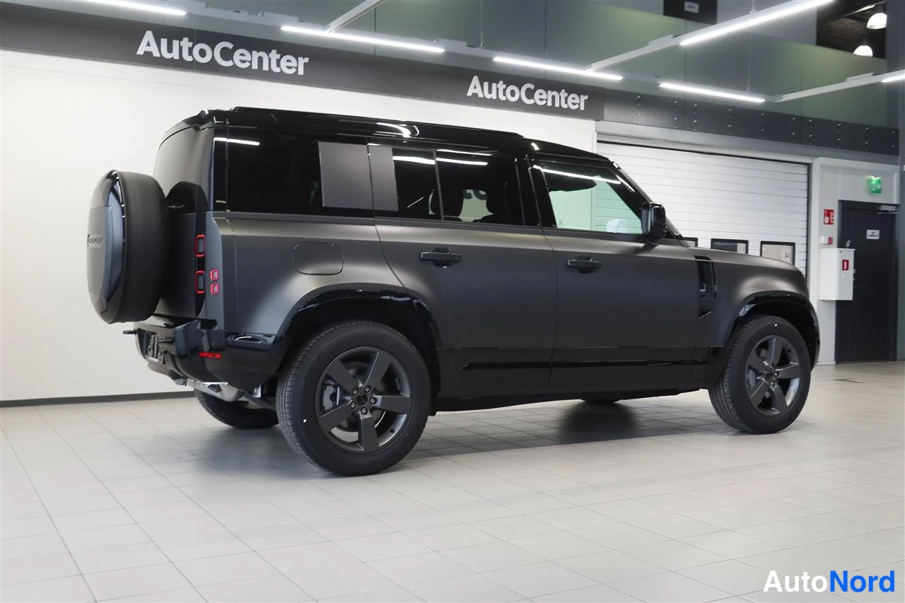 Takaviisto: Land Rover Defender sivulta-takaa kuvattuna, mattamustanharmaa kori, ulkopuolinen varapyörä takaovella, AutoCenter-hallissa.