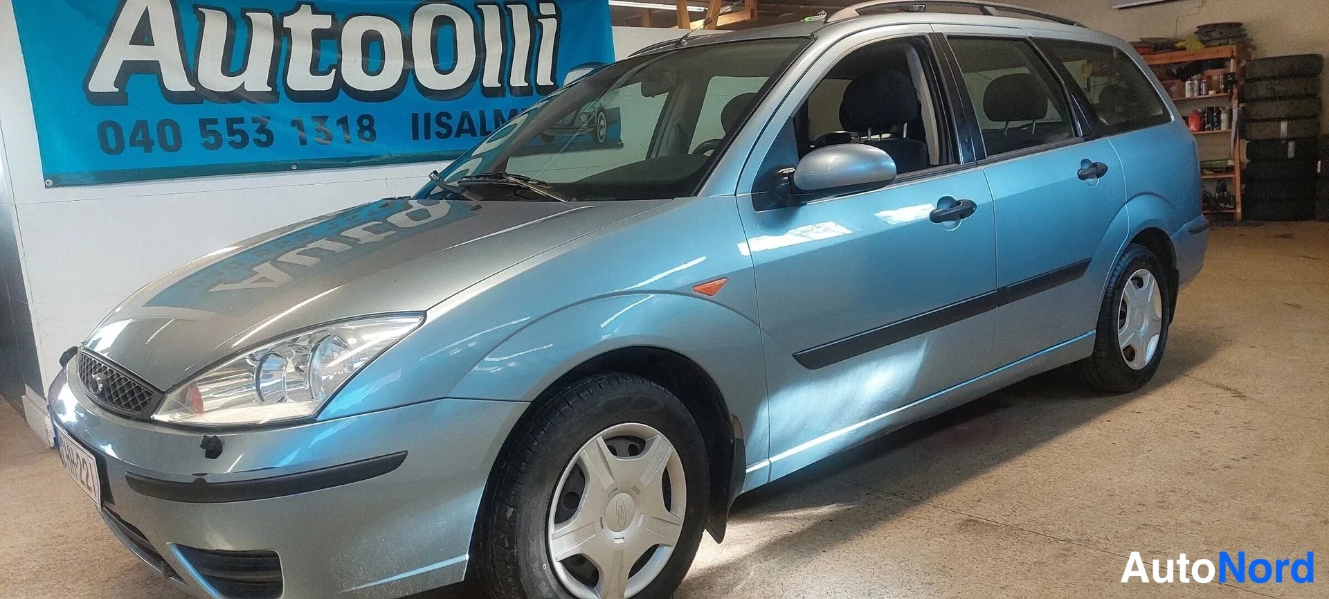 Etuviisto, nokka vasemmalle: Sinertävänharmaa farmariauto näkyvissä etukulmasta, keula vasemmalla, autotallissa Autolli-mainoksen edessä.