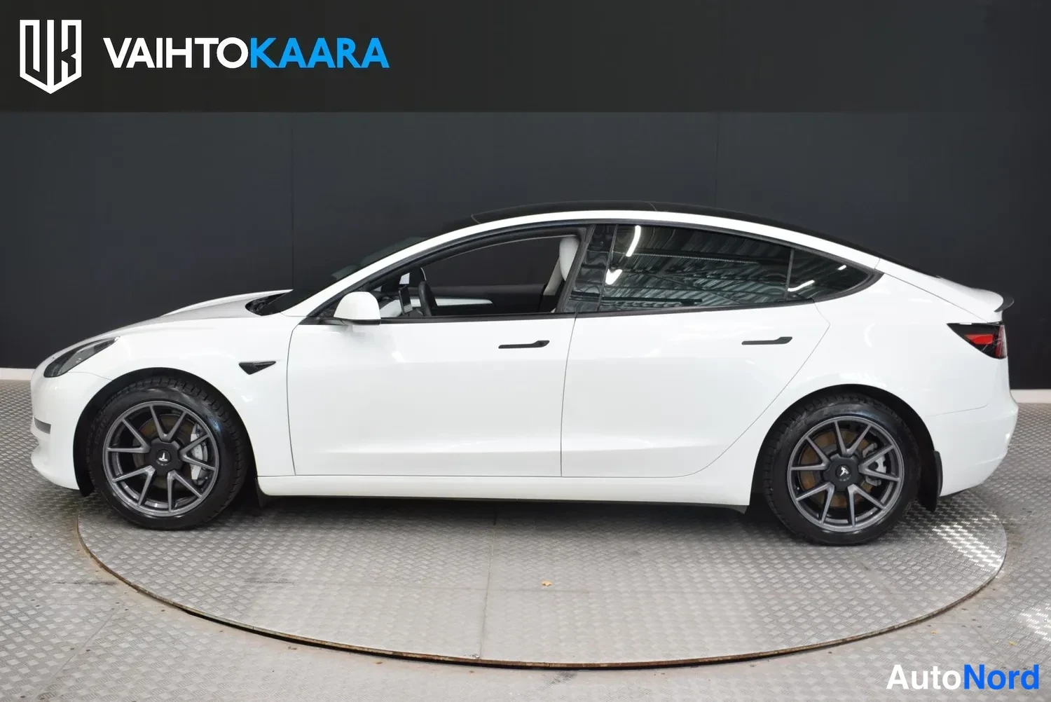 Sivulta: Tesla Model 3 -sedanin sivuprofiili valkoisena, tummat vanteet ja lasikatto studio-tilassa
