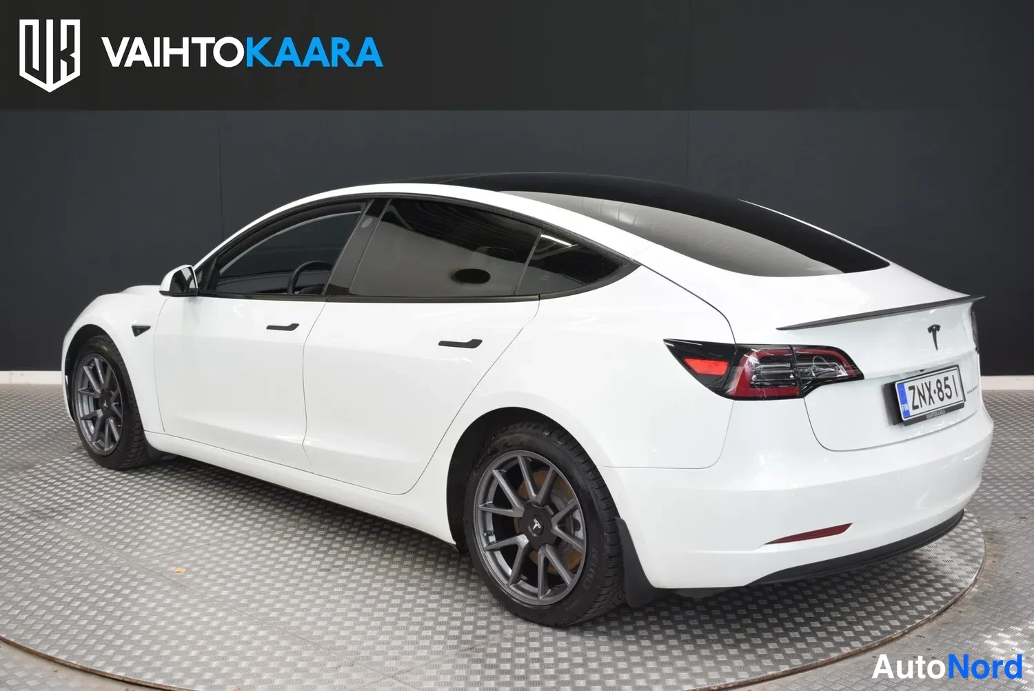 Takaviisto: Tesla Model 3 -sedanin takakulma valkoisena, rekisterikilpi ja musta spoileri
