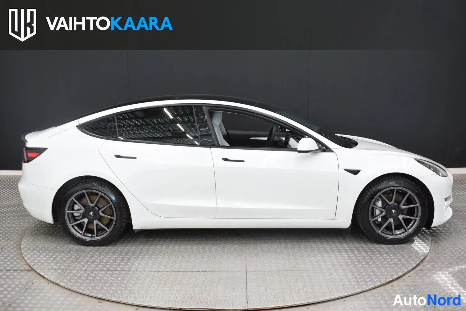 Etuviisto, nokka oikealle: Valkoinen Tesla Model 3 -sedaanin etukulmanäkymä sisätiloissa, keula oikealla.