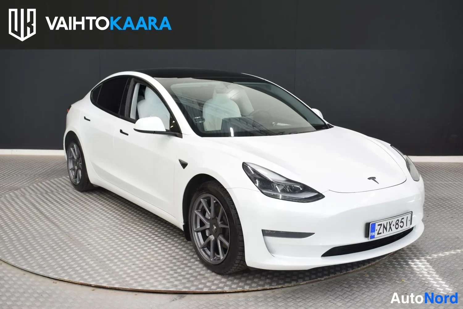 Etuviisto, nokka oikealle: Valkoinen Tesla Model 3 -sedaanin etukulmanäkymä sisätiloissa, rekisterikilpi, keula oikealla.