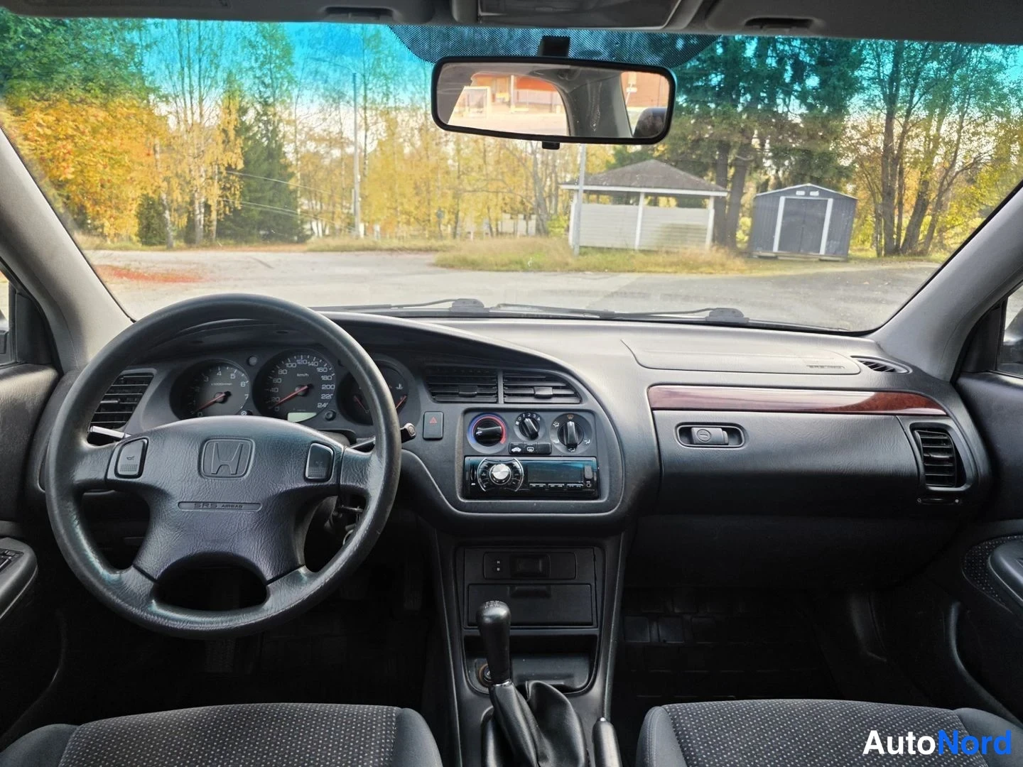 Sivulta: Näkymä kojelautaan tuulilasin läpi: Honda-ohjauspyörä, analogiset mittarit, keskellä audiopaneeli ja ilmastointisäätimet, vaihtaja manuaalivaihteistoon, tummans