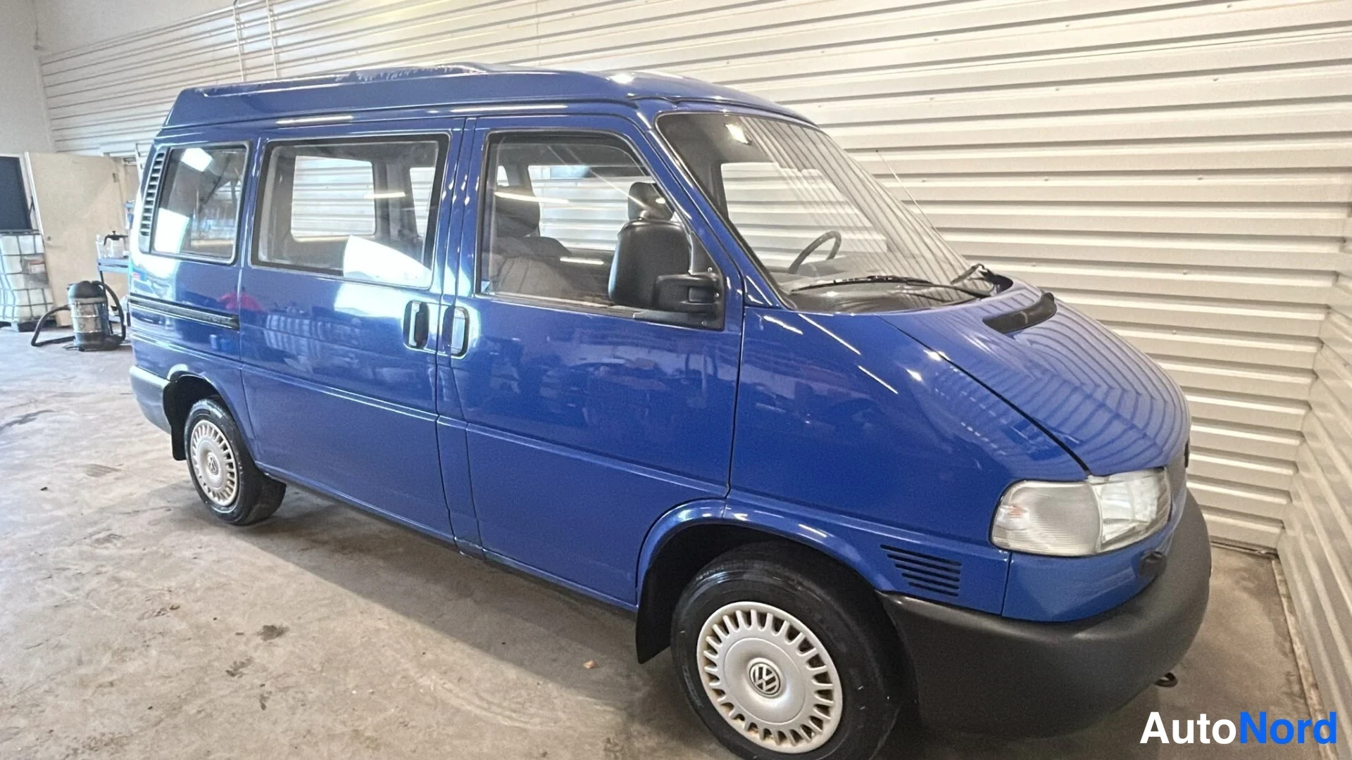 Etuviisto, nokka oikealle: Sininen Volkswagen Caravelle -pakettiauto kolmenvartalonurmakuvassa, keula oikealla, sisätiloissa valssiiniseinämäinen halli.