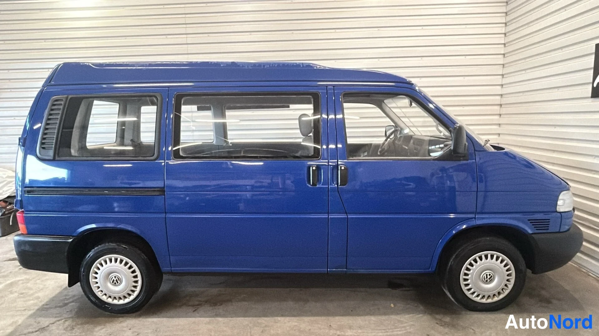Sivulta: Sinisen Volkswagen Caravelle -bussin sivuprofiili vasemmalta puolelta kuvattuna hallissa.