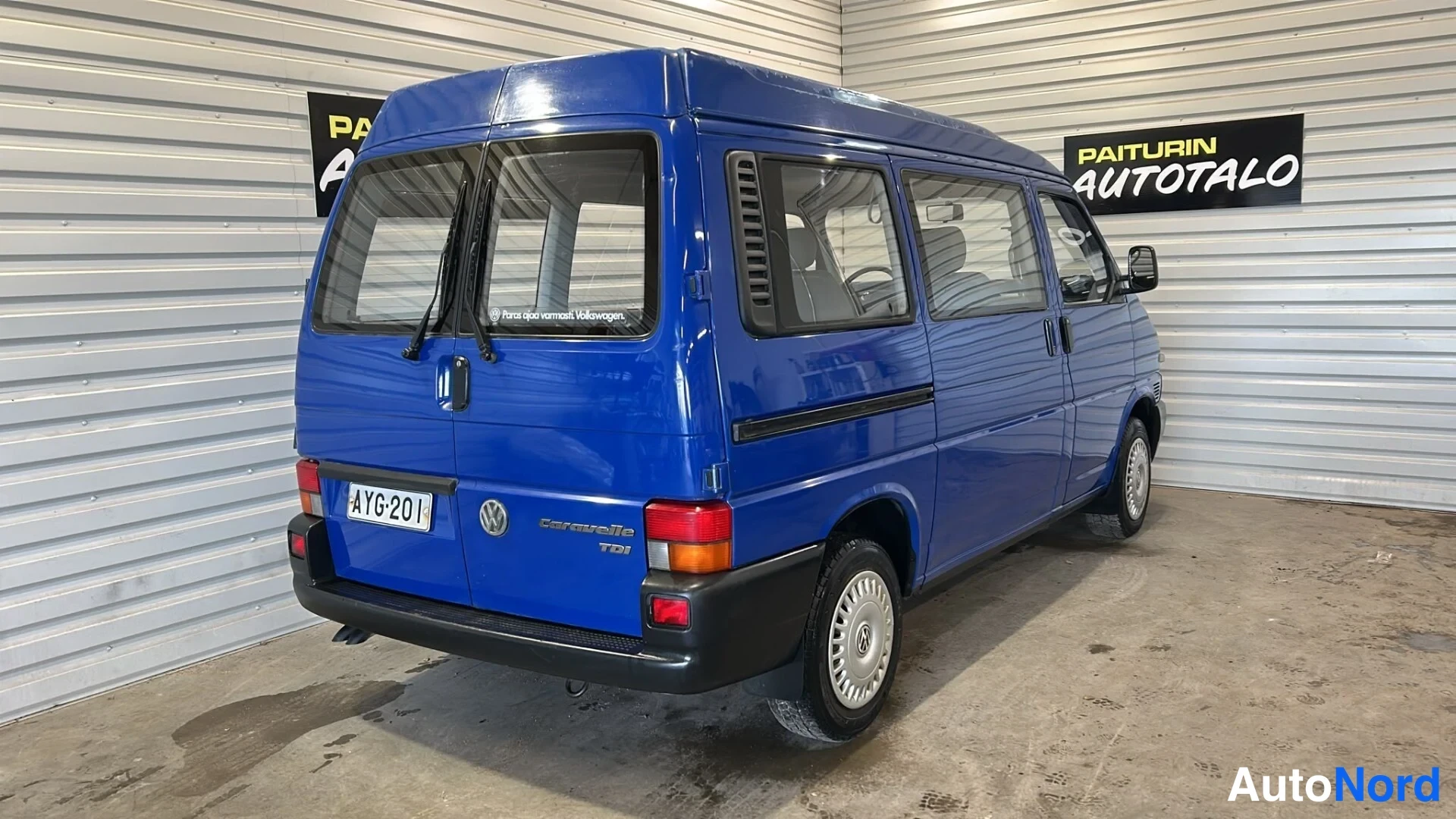 Takaviisto: Sinisen Volkswagen Caravellen takakolmannes, perä vasemmalla, rekisteritunnus, hallin seinällä Paiturin Autotalo -kyltti.