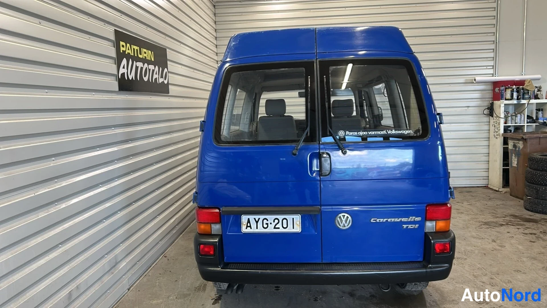 Takaa: Sinisen Volkswagen Caravellen suora takaumat, rekisteritunnus, Caravelle TDI -merkintä, Paiturin Autotalo -kyltti seinällä.